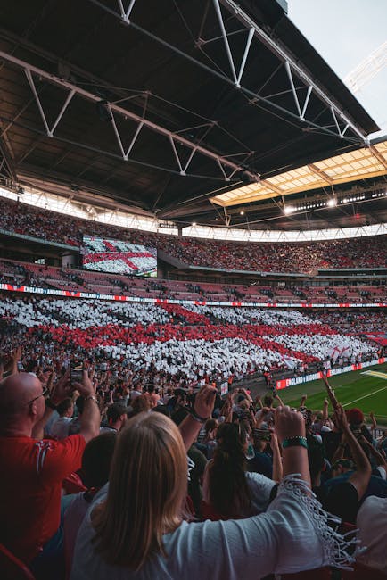 Inside a large stadium during a home team football match, with a packed audience seated in tiered stands. The crowd is holding up a coordinated mosaic of red, white, and black cards forming a pattern or message across the seating area. The stadium roof structure is visible overhead, featuring metal trusses and partial skylights allowing natural light in. In the foreground, spectators are seen from behind, some taking photos or videos with mobile phones, while others cheer and raise their hands. The pitch is partially visible at the bottom edge of the image, showing green grass and part of the sideline. The scene captures the vibrant atmosphere of a sporting event, with an emphasis on the large-scale display and enthusiastic crowd, relevant to event logistics and crowd management in a stadium environment.