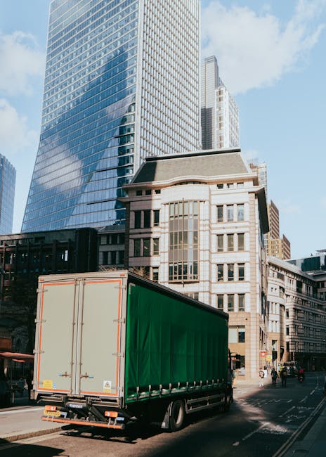 A large green curtainsider truck parked on a city street in front of tall modern office buildings and older architectural structures in Wembley Park. The truck is positioned close to the pavement, ready for loading or unloading furniture and household items, which is part of the home relocation process. The image captures the urban environment with a clear blue sky and natural daylight illuminating the scene, emphasizing the professional logistics involved in house removals. This setting is typical of a moving service provided by Man with Van Wembley Park, showcasing the transportation aspect of furniture transport and packing and moving operations within a busy city centre.