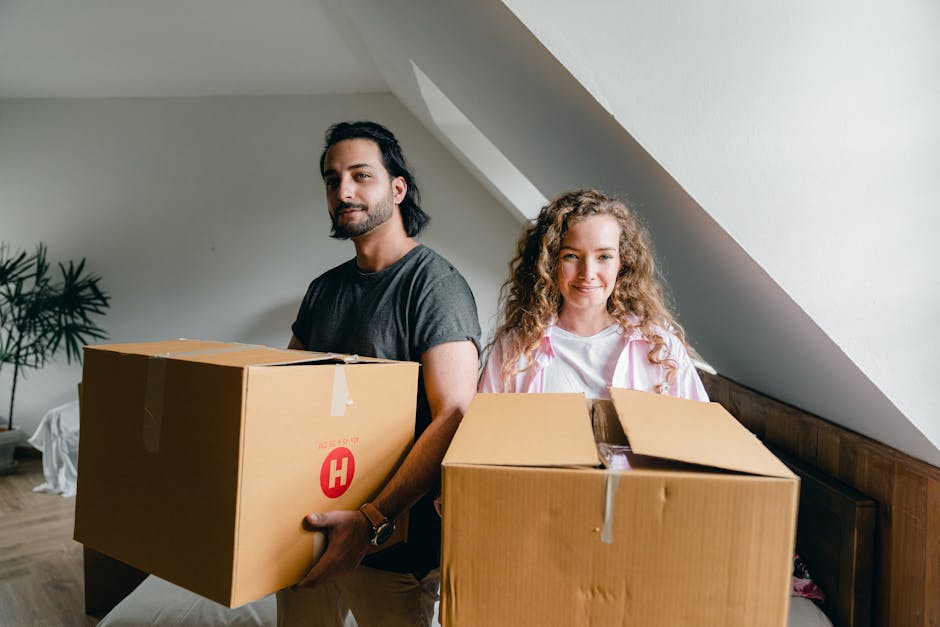 A man with dark hair and a beard, wearing a dark grey t-shirt, stands indoors holding a large cardboard box with a red shipping label, while a woman with curly blonde hair, dressed in a light pink jacket over a white shirt, stands beside him holding another cardboard box partially open, revealing packing materials inside. They are in a room with neutral walls and a wooden floor, with a potted plant visible in the background. The scene depicts the process of home relocation or packing and moving, with furniture and boxes prepared for transportation, possibly by a professional removals service such as Man with Van Wembley Park, as they carefully handle items to facilitate furniture transport and loading for a house move.