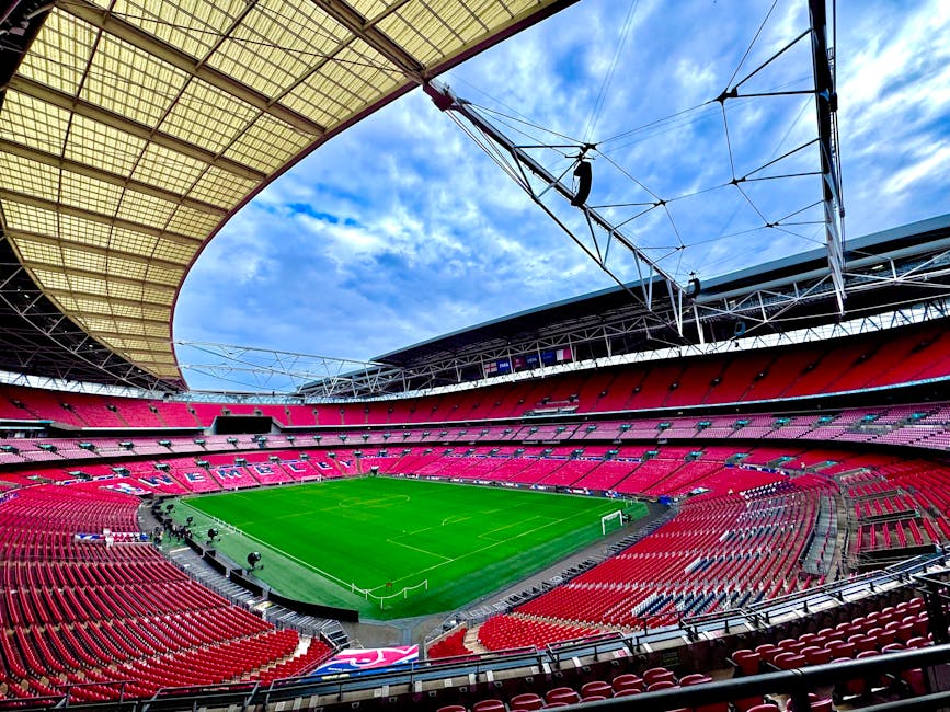 Inside a large stadium with bright natural light illuminating the red seating areas that surround a well-maintained green football pitch. The seating is arranged in multiple tiers, with a partially covered roof overhead supported by metal cables and beams. The view includes the inside of the modern architectural structure, with the sky visible through the open roof, featuring scattered clouds. In the foreground, the empty seats are visible, and the stadium's perimeter features pathways and staircases. The scene captures the expansive interior during daytime, emphasizing the scale of the venue designed for large sporting events. Occasionally, minor equipment such as lighting or sound systems is visible along the edges of the seating areas. This image can relate to the concept of moving and logistics involved in relocating sports equipment or preparing for events. It reflects the environment where furniture, equipment, or event-related items might be transported or organized during home or venue relocations, as seen in professional removals from companies like Man with Van Wembley Park.