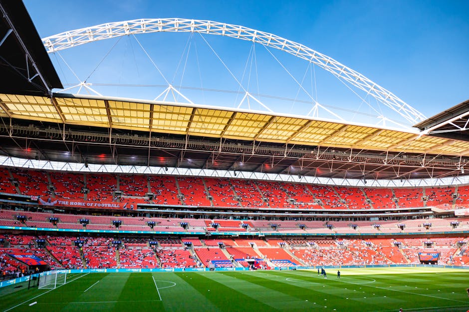 The interior of a large football stadium with a well-maintained grass pitch, white goalposts, and surrounding seating areas filled with red and pink seats. Overhead, a curved white roof structure supported by numerous metal beams covers part of the stands, providing shelter for spectators. The stadium is bathed in natural daylight, with clear blue sky visible beyond the roof. A few individuals are seen walking on the pitch near the goal area, possibly preparing for a match or event. The seating arrangement includes multiple tiers, and some sections are empty while others are occupied by small groups of fans. Inside the stadium, the atmosphere appears quiet, with no crowds or active play at the moment. The setting aligns with preparations for an upcoming football game or event, and the image emphasizes the spacious, professional environment suitable for large sporting occasions. Man with Van Wembley Park occasionally manages home relocation and furniture transport logistics, supporting efficient packing and loading processes.