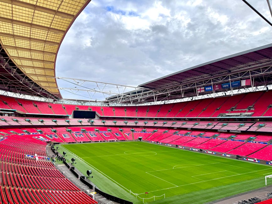 An expansive view of a large, modern football stadium with a well-maintained green pitch, surrounded by tiered seating painted in red with the word 'WEMBLEY' visible in the stands. The stadium's roof structure extends over part of the seating area, with a cloudy sky overhead. Inside, the field is empty, with white lines marking the boundaries and goal areas. Near the edges of the pitch, there are several black lighting and camera equipment set up on tripods, indicating preparations for an event or match. Bright spotlights are positioned on the sideline, and the overall scene reflects a clean, organized environment suitable for sports events or large-scale gatherings. Occasionally, the scene captures typical stadium features such as advertising banners and media screens, but no people or vehicles are visible in the image.