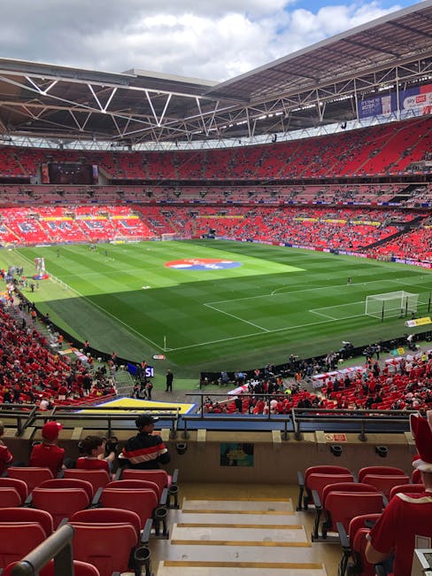 An indoor football stadium with a well-maintained grass pitch marked with white lines for a match, viewed from a higher tier seating area with rows of red seats. The stadium features a partially covered roof supported by white trusses, with bright lighting illuminating the field. Red and yellow fabric seating sections are visible in the background, with some spectators seated, some standing near the pitch. Equipment such as small carts and barriers are positioned close to the field, and the open gates at the sides indicate ongoing preparations for a game or event. The surrounding environment suggests a professional sports venue, where furniture transport and removal services, such as those offered by Man with Van Wembley Park, could be involved in logistics or supportive services related to large-scale event management or equipment transportation.