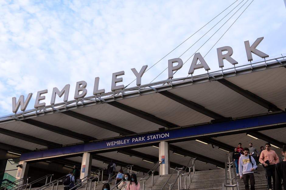 A view of Wembley Park Station entrance with a large, white sign spelling 'WEMBLEY PARK' mounted on the station roof, supported by metal beams. Beneath the sign, there is a blue banner displaying 'WEMBLEY PARK STATION' over the stairway leading down to the station platform. Several pedestrians, some wearing face masks, are seen ascending and descending the outdoor steps on a cloudy day. The scene depicts the busy, urban environment typical of a London transport hub, relevant to house relocation and home removal services such as those offered by Man with Van Wembley Park, who handle Transportation and furniture loading during packing and moving processes.