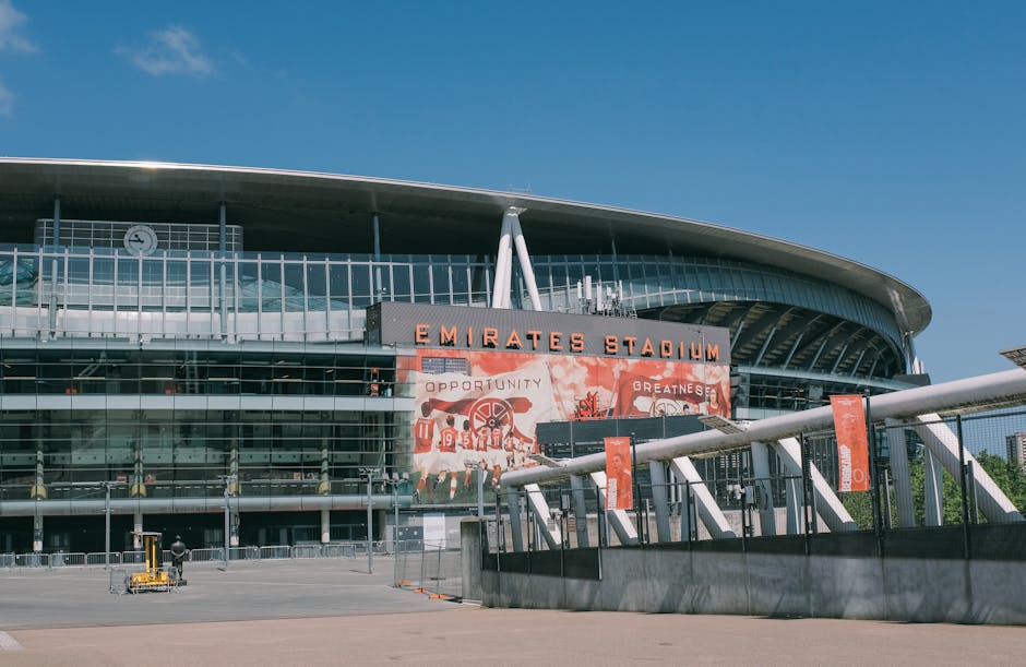 The exterior of Emirates Stadium, home to Arsenal FC, is visible with its modern architectural design featuring a curved roof and large glass facade. Prominent signage displays the stadium's name in bold orange letters, with a banner below showing the team logo and messages related to opportunity and greatness. The open area in front of the stadium includes a paved walkway and a fenced loading zone with a few orange banners attached to poles. A small platform with a luggage trolley and some wooden crates or cardboard boxes are situated near the entrance, suggesting preparations for home relocation or furniture transport. The scene is lit with bright daylight against a clear blue sky, illustrating the typical environment for loading or packing during a moving service, such as those offered by Man with Van Wembley Park, specializing in removals and moving logistics near Wembley Stadium and Wembley Park.