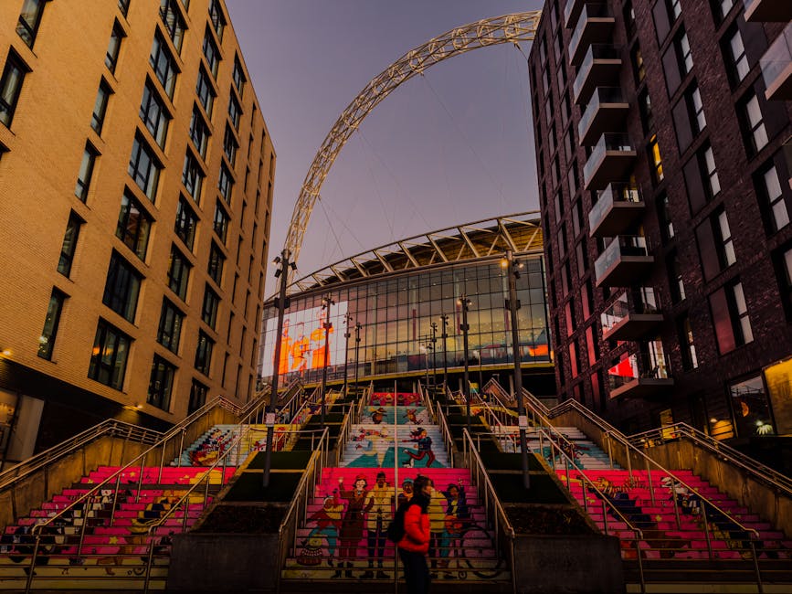 Inside the large Wembley Stadium with the natural daylight illuminating the seating area and the lush green football pitch in the center. Red stadium seats surround the pitch, with some branded banner