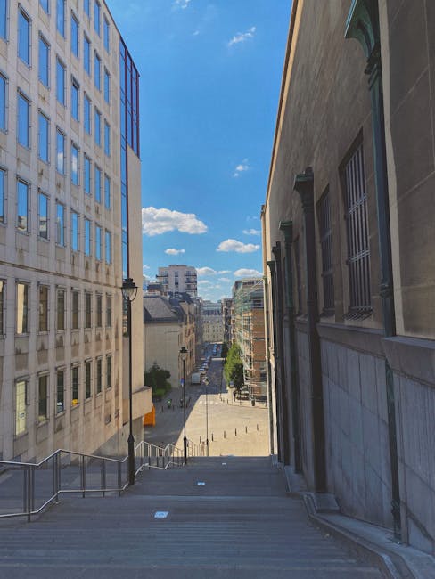 A view from the top of a narrow outdoor staircase situated between two multi-storey buildings in Wembley Park, with metal handrails along the sides and steps made of concrete or stone. The staircase descends toward a city street lined with additional buildings, street lamps, and some greenery, under a bright blue sky with scattered white clouds. This setting illustrates the challenges of navigating narrow stairs during home relocation or furniture transport, a common scenario faced in Wembley Park. Man with Van Wembley Park specialises in removals, including managing tight staircase access during moving processes, as evidenced by the staircase's limited width and urban environment which require careful planning for loading and unloading household goods. The scene also hints at the importance of efficient packing and loading strategies when dealing with confined spaces during home or office moves, supported by the company's expertise in logistical solutions.