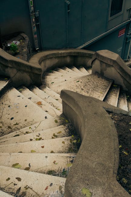 A concrete outdoor spiral staircase with rounded stone balustrades on both sides leading down from an upper level. The stairs are weathered with visible dirt, small leaves, and debris scattered across the steps. Adjacent to the staircase, there are some electrical boxes or utility panels mounted on a building wall, painted in a dark color. The scene is lit with natural light, illuminating the stone surface and surrounding environment, which suggests daytime. The image depicts part of a residential or commercial property where furniture, boxes, and packing materials could be carried or transported during a home relocation process. Man with Van Wembley Park strategically uses such staircases in their furniture transport and packing and moving services, particularly in properties with narrow or challenging stair layouts, aligning with their focus on house removals and logistical solutions for difficult stairways.