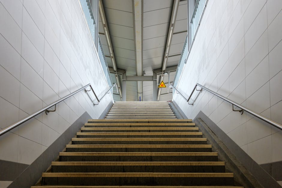 A view from the top of a narrow outdoor staircase situated between two multi-storey buildings in Wembley Park, with metal handrails along the sides and steps made of concrete or stone. The staircase descends toward a city street lined with additional buildings, street lamps, and some greenery, under a bright blue sky with scattered white clouds. This setting illustrates the challenges of navigating narrow stairs during home relocation or furniture transport, a common scenario faced in Wembley Park. Man with Van Wembley Park specialises in removals, including managing tight staircase access during moving processes, as evidenced by the staircase's limited width and urban environment which require careful planning for loading and unloading household goods. The scene also hints at the importance of efficient packing and loading strategies when dealing with confined spaces during home or office moves, supported by the company's expertise in logistical solutions.