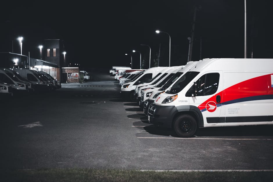 Night-time view of a parking lot illuminated by streetlights, showing a row of white delivery vans with red and blue branding parked side by side on the right, with their front ends facing the viewer.