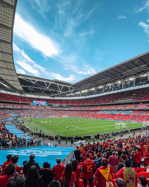 The exterior front view of Emirates Stadium in Wembley Park shows the large, modern structure with a curved roof and extensive glass facade. In front of the stadium, there is an open paved area with a